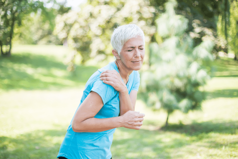 The difference between soreness and pain is often dependent on the cause: a senior woman rubs her achy shoulder outdoors.