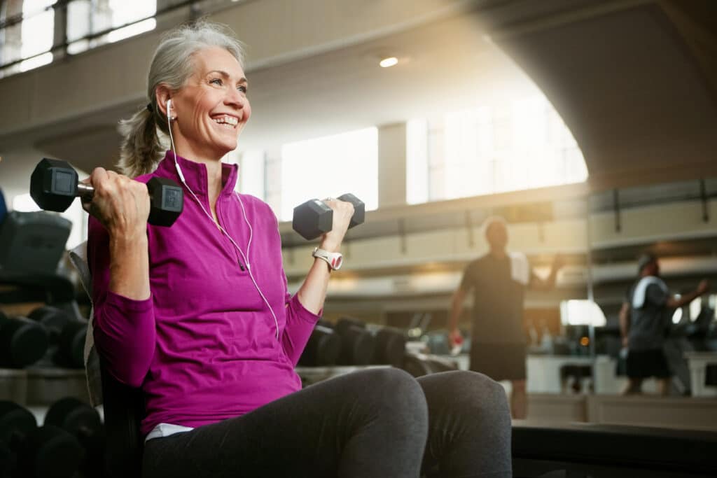 Improving bone health before surgery: an older woman lifts weights at the gym, smiling.
