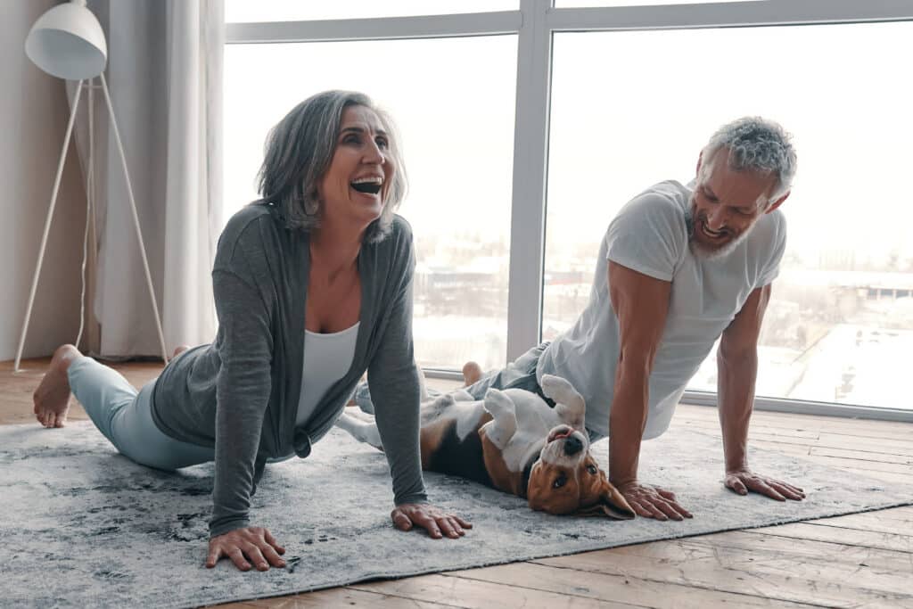 Image of senior couple doing yoga stretches on the floor with their dog.  Exercise can improve mood and other cognitive health attributes.
