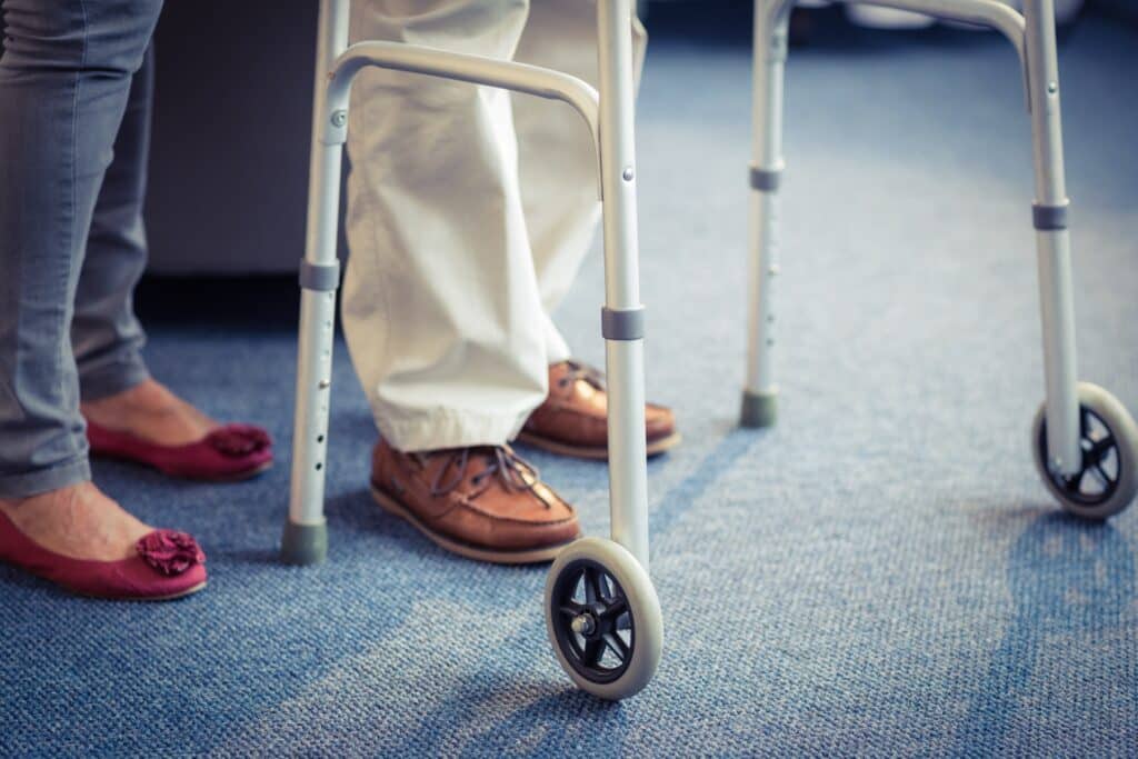 Image of wheeled walker being used by a senior man with a woman behind him (only feet are visible)