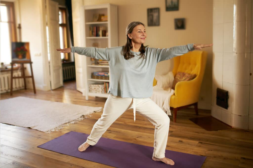 Image of a smiling older woman doing a warrior II poses to improve hip rotation for injury prevention.