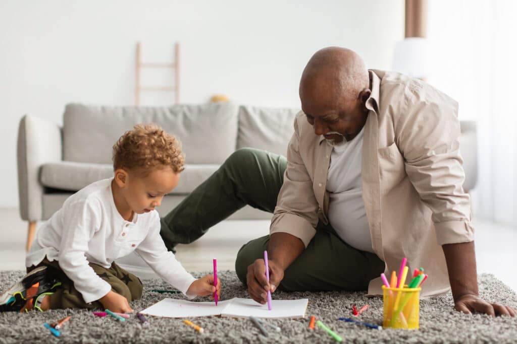 African American grandfather drawing on the floor with his grandson.  Getting up and down from the floor is one of the functional movements that is much easier when focusing on using proper body mechanics.