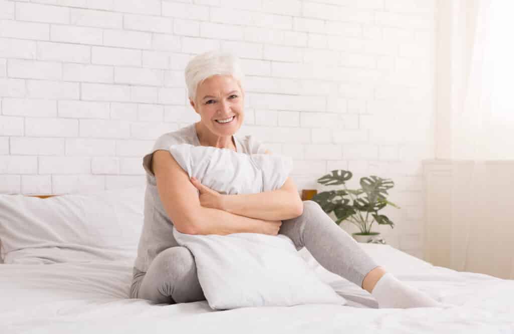 Image of a happy older woman sitting in bed holding a pillow.  A pillow can be a helpful tool for managing pain while asleep