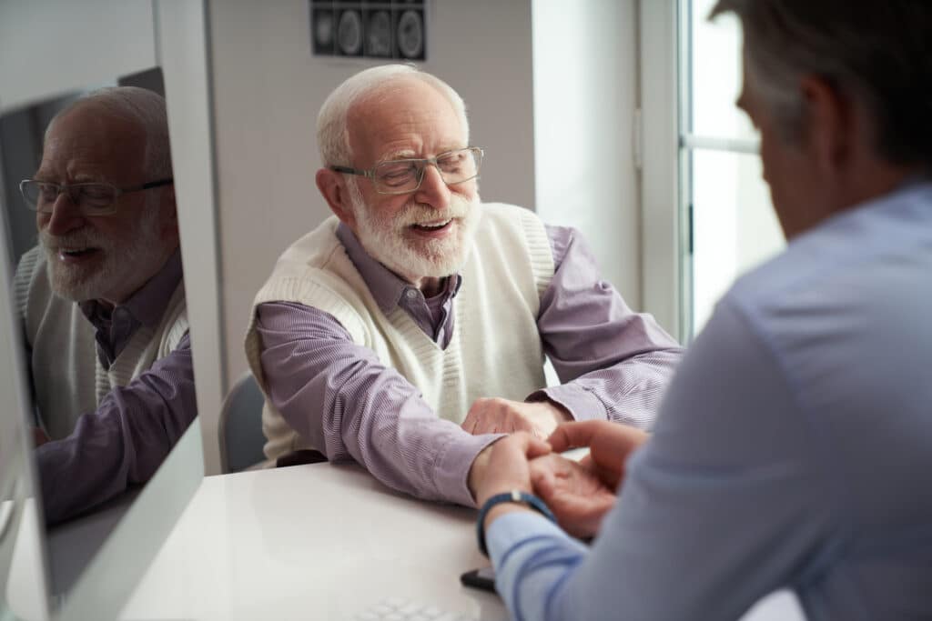 Elderly man smiling while sitting on a chair having his hand extended to a doctor checking for deviations