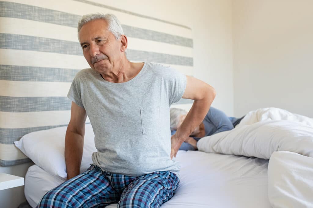 Image of a senior man with hip pain while sleeping, awake at the edge of the bed with his wife.