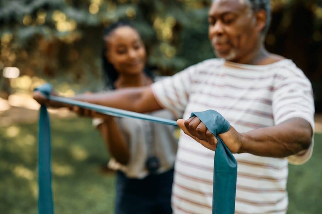 Close up of black senior using resistance band during physical therapy at nursing home's park.