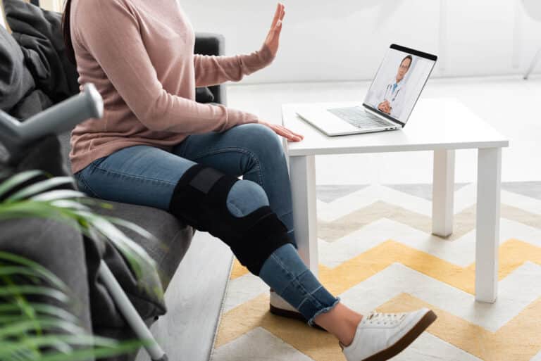 Choosing the right knee brace: image of a woman wearing a knee brace while sitting on the couch having a zoom call with her doctor.