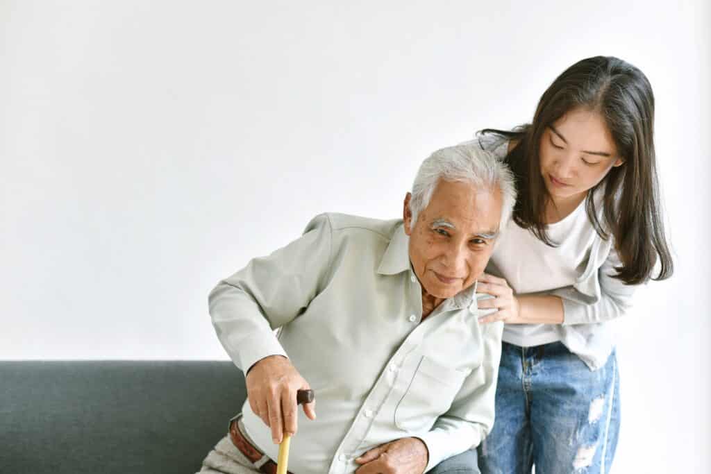 Image of an older man being assisted by his daughter while getting up from a couch using a cane.