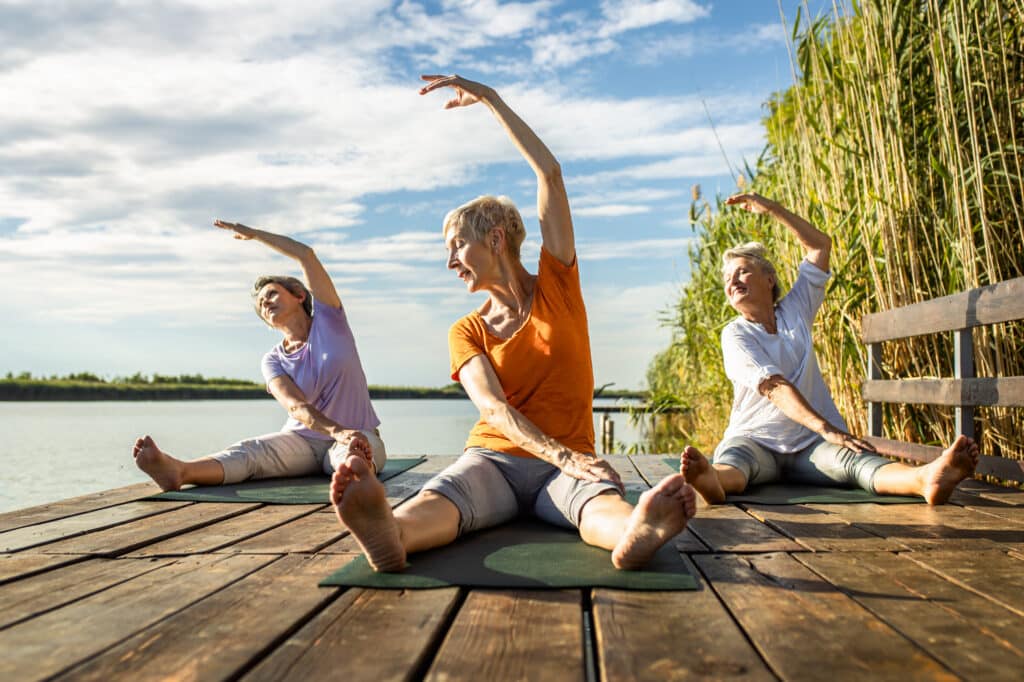 Three senior women doing yoga on a dock in the morning.