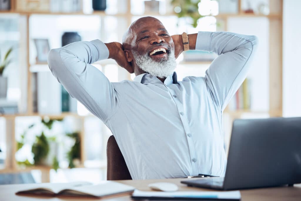 Laughing older black man seated at his work desk stretching his back to incorporate exercise into his work routine.