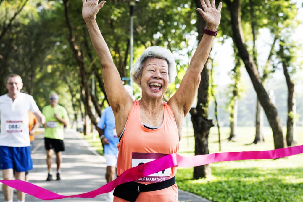 Image of a healthy senior Asian woman crossing the finish line in a running race.  Consistent daily exercise is key to longevity, and it's a marathon, not a sprint.