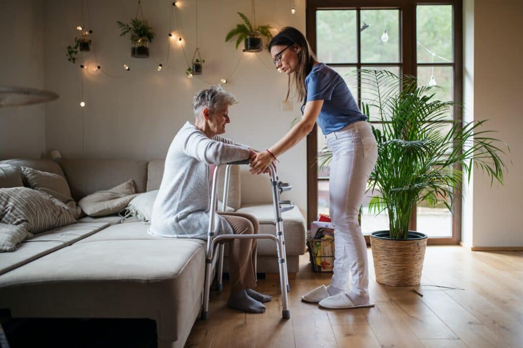 Healthcare worker or caregiver visiting senior woman indoors at home, helping her to walk.