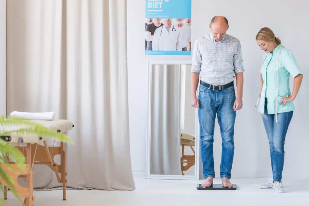 An older man weighs himself on a scale beside his wife