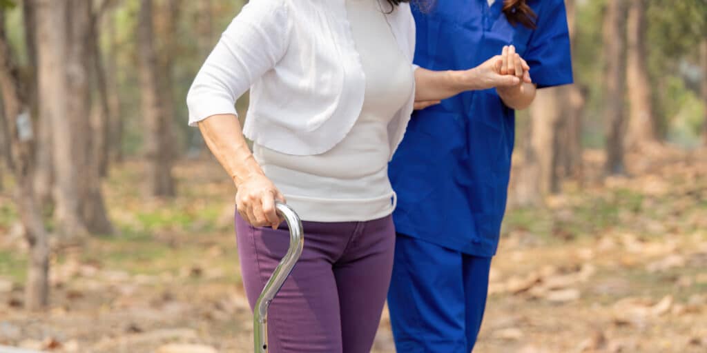 How to do a balance test at home: a woman with a cane gets assistance from a nurse while walking in the woods.