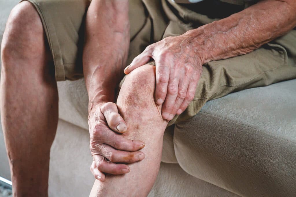 An older man holds his achy knees while sitting on the couch