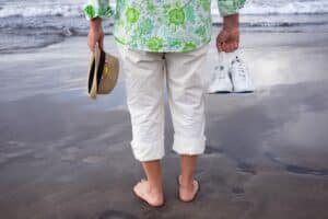 Rear view of senior man barefoot walking on sea shore holding shoes and hat in hands