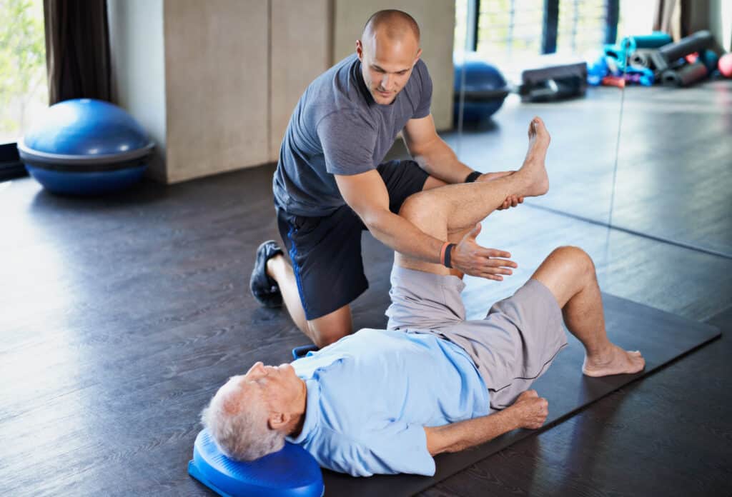 MASH treatment: a physical therapist evaluates a senior patient's hip pain while he lies on the floor.