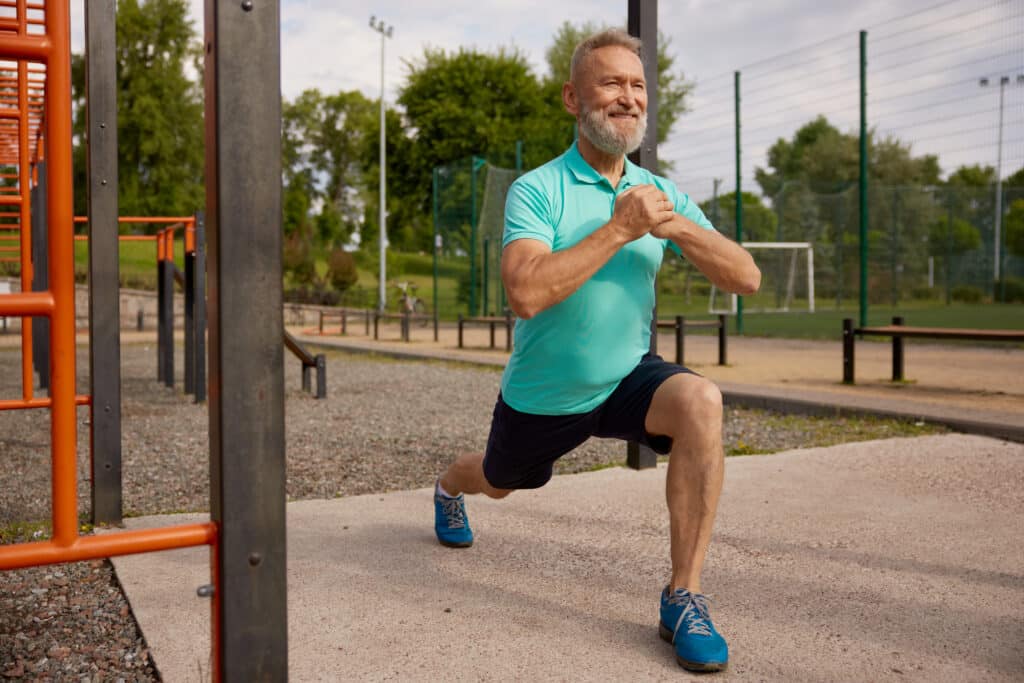 An older man does a lunge hip extension stretch at the park.