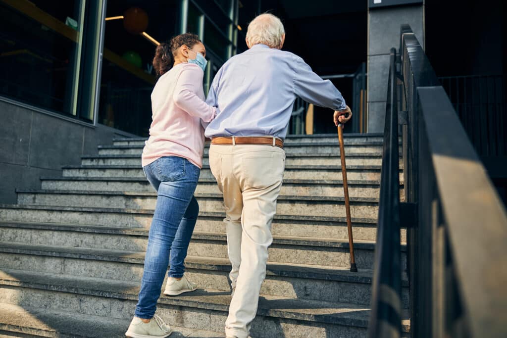 Younger woman helping older man with a cane as he walks up stairs outdoors